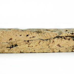 May include: Close-up of a cross-section of a cork board. The cork has a light brown color with a rough, textured surface. The layers of the cork are visible, with some cracks and holes throughout. The background is white.