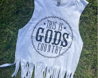 May include: White sleeveless shirt with a frayed hem. The shirt has a black graphic print that says "This is God's Country" inside a barbed wire circle.