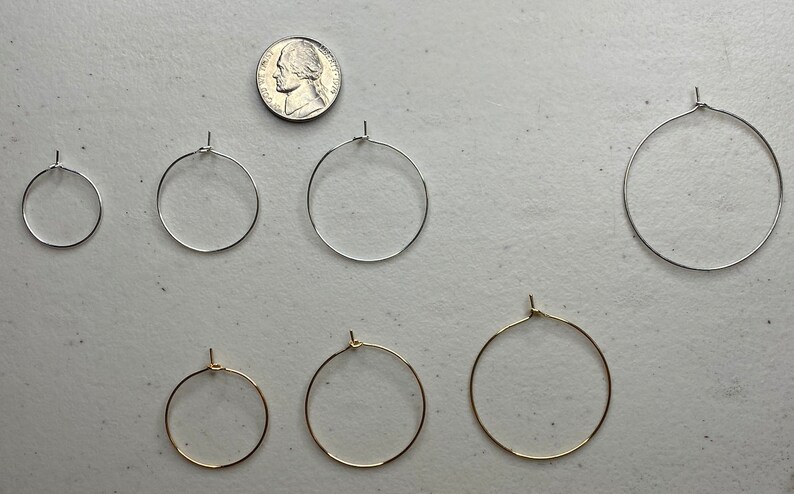 Puede incluir: Una variedad de pendientes de aro en tonos plateados y dorados, exhibidos sobre una superficie clara. Se incluye una moneda para mostrar la escala. Los pendientes tienen un dise&ntilde;o simple y cl&aacute;sico, adecuados para el uso diario. Medidas en cent&iacute;metros.