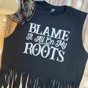 K&ouml;nnte beinhalten: Schwarzes &auml;rmelloses Shirt mit einem wei&szlig;en Aufdruck, der "Blame It All On My Roots" mit zwei Sternen sagt. Das Shirt hat einen Fransen-Saum.
