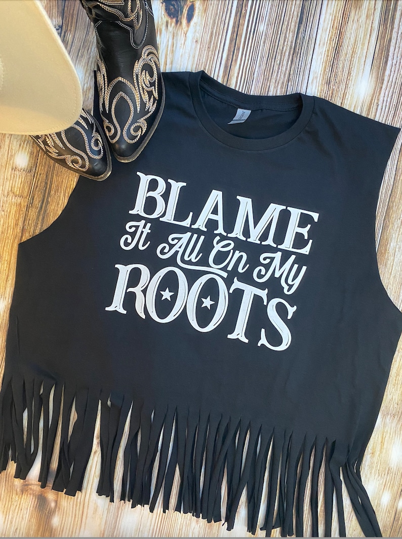 K&ouml;nnte beinhalten: Schwarzes &auml;rmelloses Shirt mit einem wei&szlig;en Aufdruck, der "Blame It All On My Roots" mit zwei Sternen sagt. Das Shirt hat einen Fransen-Saum.