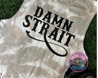 May include: A white tie-dye sleeveless shirt with the text "DAMN STRAIT" in black lettering. The shirt has a frayed hem and is paired with a pair of black cowboy boots. The image also includes a small logo for "Backstage Customs" with a guitar icon.