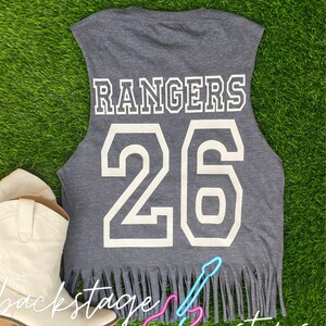 Pu&ograve; includere: Maglietta grigia senza maniche con bordi sfrangiati. La parte posteriore della maglietta ha la parola "RANGERS" e il numero "26" stampati in bianco. La maglietta ha una grafica di chitarra con il testo "backstage customs" stampato sotto.
