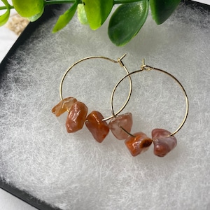 Puede incluir: Pendientes de aro dorados con cuentas de piedras preciosas de color naranja y beige. Los pendientes se presentan en una caja negra con relleno de algod&oacute;n blanco, con hojas verdes en el fondo. Los pendientes tienen un dise&ntilde;o sencillo y elegante.