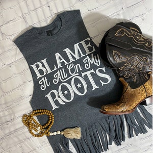 K&ouml;nnte beinhalten: Graues &auml;rmelloses Shirt mit einem ausgefransten Saum. Das Shirt hat wei&szlig;en Text, der "BLAME It All On My ROOTS" mit zwei Sternen &uuml;ber dem Wort "ROOTS" lautet.
