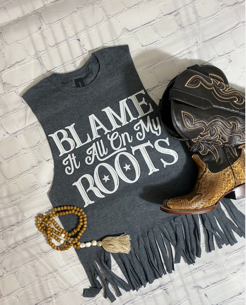 K&ouml;nnte beinhalten: Graues &auml;rmelloses Shirt mit einem ausgefransten Saum. Das Shirt hat wei&szlig;en Text, der "BLAME It All On My ROOTS" mit zwei Sternen &uuml;ber dem Wort "ROOTS" lautet.