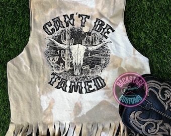 Könnte beinhalten: Ein weißes ärmelloses Shirt mit Batik-Muster und Fransenbesatz. Das Shirt zeigt eine schwarze Grafik eines Schädels mit Hörnern und dem Text "Can't Be Tamed".