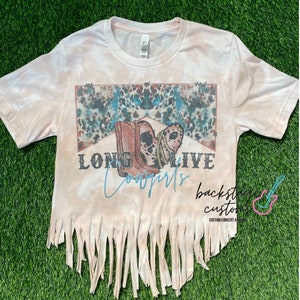 May include: A light pink tie-dye t-shirt with a graphic of cowboy boots and the words "LONG LIVE Cowgirls". The bottom hem is fringed. The shirt is on a green surface.