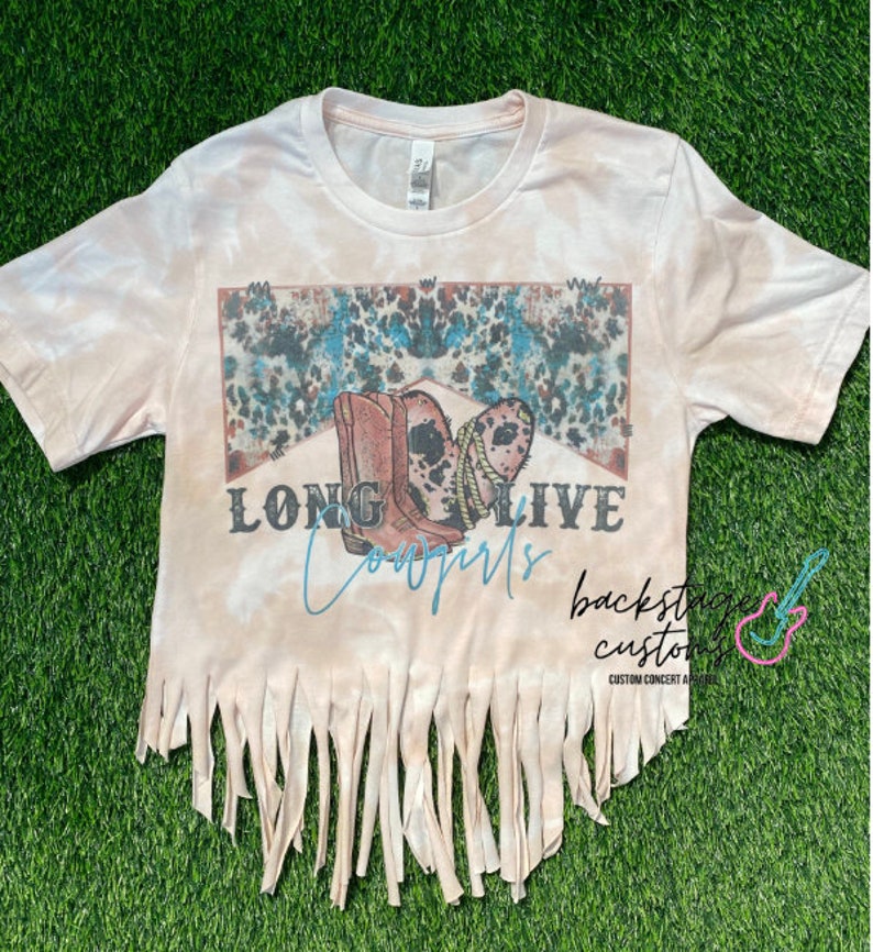 May include: A light pink tie-dye t-shirt with a graphic of cowboy boots and the words "LONG LIVE Cowgirls". The bottom hem is fringed. The shirt is on a green surface.