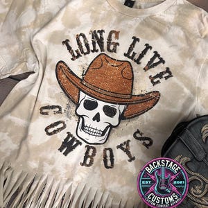 May include: A beige tie-dye t-shirt with a distressed graphic of a skull wearing a cowboy hat and the text "Long Live Cowboys". The shirt has a frayed hem.