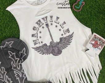 Op de afbeelding: Een wit, mouwloos shirt met een distressed look en franjes. Het shirt heeft een zwart-witte afbeelding van een gitaar met vleugels en het woord "Nashville" in een cirkel eromheen. Het shirt ligt op een groene grasmat met een paar zwarte cowboylaarzen.