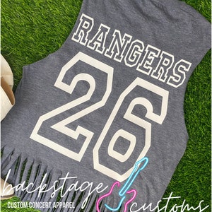 Pu&ograve; includere: Maglietta grigia senza maniche con la parola "RANGERS" e il numero "26" stampati in bianco sul retro. La maglietta ha un orlo a frangia e il testo "backstage customs" stampato sul fondo.