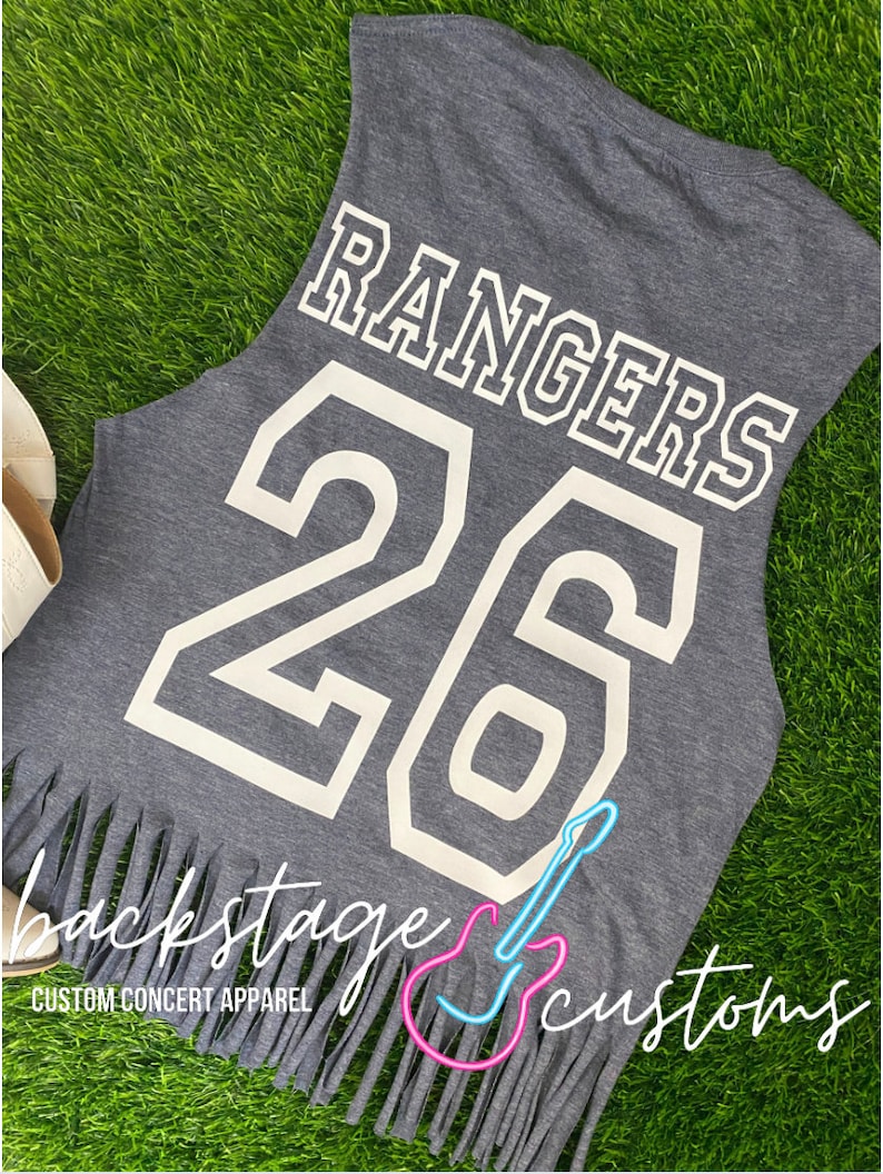 Pu&ograve; includere: Maglietta grigia senza maniche con la parola "RANGERS" e il numero "26" stampati in bianco sul retro. La maglietta ha un orlo a frangia e il testo "backstage customs" stampato sul fondo.