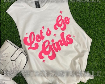 May include: White sleeveless shirt with the text "Let's go girls" in pink script with stars. The shirt is laying on a green grassy surface with white cowboy boots.
