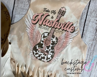 May include: A light brown sleeveless shirt with a tie-dye pattern and fringe trim. The shirt features a graphic of a guitar with wings and the text "Take Me to Nashville Music City". The text is in pink and brown with glitter accents. The shirt is from Backstage Customs, a custom concert apparel company.