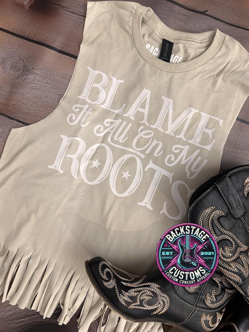 K&ouml;nnte beinhalten: Ein beiges &auml;rmelloses Shirt mit einem wei&szlig;en Aufdruck, der "Blame It All On My Roots" sagt, und einem Fransenbesatz. Das Shirt wird mit einem schwarzen Cowboy-Stiefel mit Strasssteinen kombiniert.