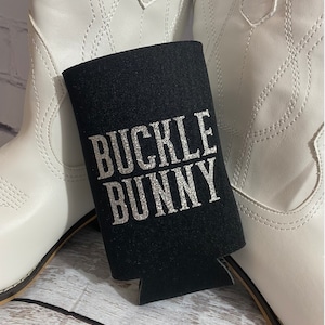 May include: Black can cooler with the text "BUCKLE BUNNY" in silver glitter lettering. The cooler is sitting on a wooden surface next to a pair of white cowboy boots.