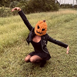 Puede incluir: Una persona con una chaqueta de cuero negra y un vestido negro está agachada en un campo de hierba. Lleva una máscara de calabaza de ganchillo con una cara de miedo.
