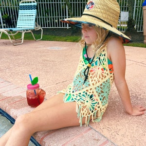 Puede incluir: Una persona joven lleva una cubierta de crochet amarilla y verde con flecos, un sombrero de paja con un diseño colorido y un collar negro se sienta junto a una piscina con un tarro de cristal rosa y verde lleno de una bebida roja y una pajita azul.