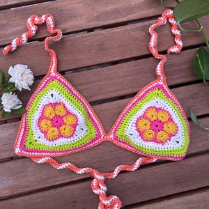 Puede incluir: Top de bikini de ganchillo hecho a mano con un diseño floral. El top presenta una vibrante paleta de colores rosa, amarillo, verde y blanco, con correas coral y blancas. Las copas triangulares están adornadas con un motivo floral.