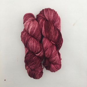 Può includere: Due gomitoli di filato rosso scuro con una sottile variazione di colore bianco. Il filato è attorcigliato in un nodo.