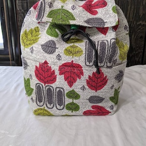 May include: A white drawstring backpack with a leaf pattern in shades of green, red, and gray. The backpack has a green drawstring and a flap with a fold-over closure.