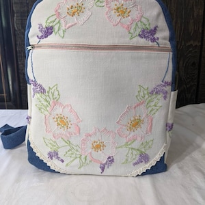 May include: A white canvas backpack with a blue denim trim and pink and purple embroidered flowers. The backpack has a zipper closure and a small pocket on the side.