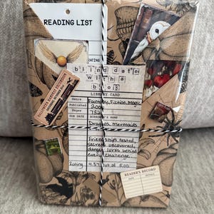 May include: A book wrapped in brown paper with a black and white botanical and insect print, tied with black and white twine. It features a library card with the text "blind date with a book" and "READING LIST," plus stickers of an owl and a golden snitch.