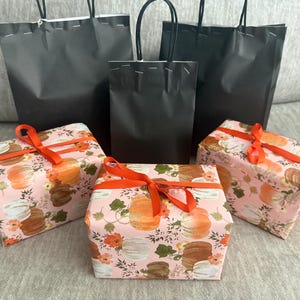 May include: Three rectangular gift boxes wrapped in light pink paper featuring a pattern of orange, white, and brown pumpkins with green leaves, tied with orange ribbons, positioned in front of three plain black paper shopping bags with handles.