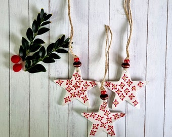 May include: Three white ceramic star ornaments with red floral patterns and twine hangers. The stars are hanging against a white wood background.