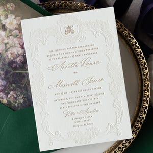 以下が含まれることがあります： エンボス加工されたフローラルディテールと金色の文字が特徴のエレガントな結婚式の招待状。招待状には、アネット・ラウラとマクスウェル・ジェームズの名前、結婚式の日付と場所が記載されています。招待状は白いカードにあり、金色の装飾フレームが付いています。