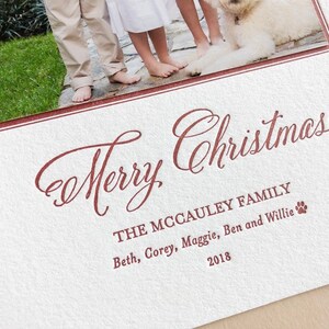 May include: A white textured holiday card featuring a photograph of a family and a dog at the top. The text Merry Christmas is printed in a dark red script font, followed by The McCauley Family, the names Beth, Corey, Maggie, Ben and Willie, and 2018.