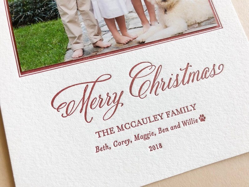 May include: A white textured holiday card featuring a photograph of a family and a dog at the top. The text Merry Christmas is printed in a dark red script font, followed by The McCauley Family, the names Beth, Corey, Maggie, Ben and Willie, and 2018.