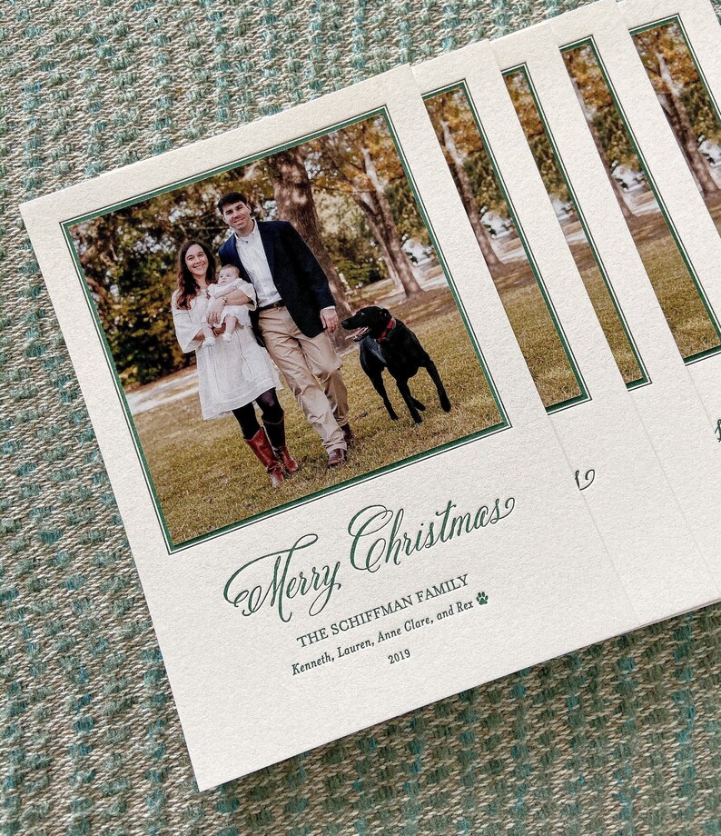 May include: A stack of rectangular holiday cards featuring a photograph of two adults, an infant, and a black dog outdoors. The text reads Merry Christmas, THE SCHIFFMAN FAMILY, Kenneth, Lauren, Anne Clare, and Rex, and the year 2019.