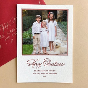 May include: A rectangular white Christmas card featuring a photograph of three children and a white dog on a stone path. The card includes the text Merry Christmas, The McCauley Family, Beth, Corey, Maggie, Ben and Willie, and 2018 in red ink.
