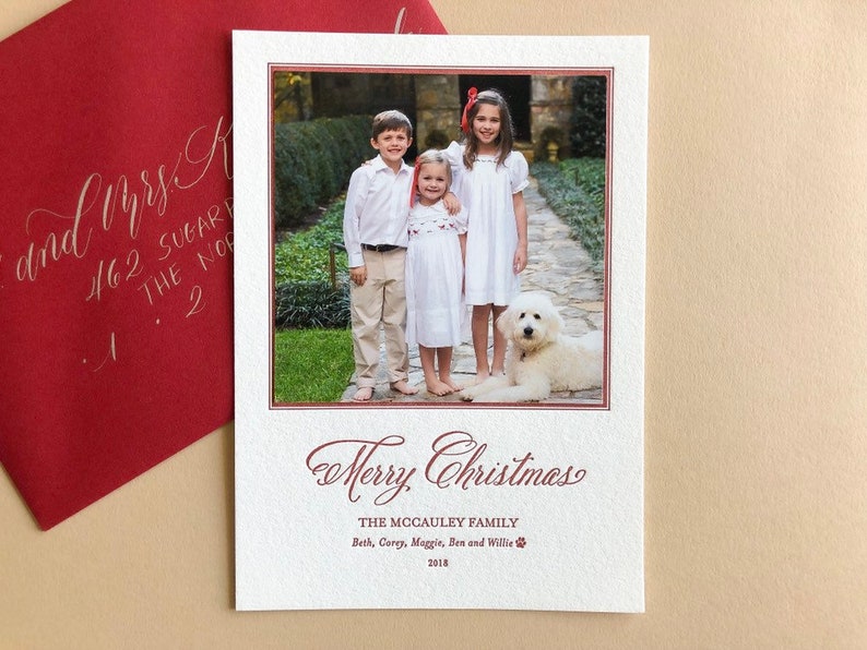 May include: A rectangular white Christmas card featuring a photograph of three children and a white dog on a stone path. The card includes the text Merry Christmas, The McCauley Family, Beth, Corey, Maggie, Ben and Willie, and 2018 in red ink.