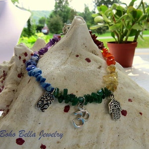 May include: A rainbow chakra bracelet with silver charms, including a hamsa hand and an om symbol. The bracelet is made with natural stones and is displayed on a seashell.