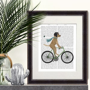 以下が含まれることがあります： 緑色の自転車に乗った犬が描かれた額入りのアートプリント。犬は青いスカーフと黒い飛行士キャップを着用しています。アートワークは、ヴィンテージの書籍ページを背景に、濃い茶色のフレームに入っています。