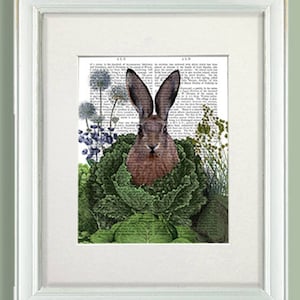 May include: A framed print of a brown rabbit sitting in a bed of green cabbage and other plants. The print is on a white background and has a white frame.