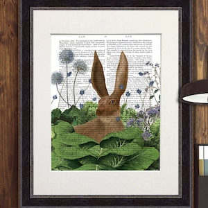 Op de afbeelding: Ingelijste kunstdruk met een bruine konijnenillustratie. Het konijn staat tegen een achtergrond van vintage boekpagina's, omringd door groene bladeren en blauwe bloemen. Het kunstwerk zit in een zwarte lijst met een passe-partout.