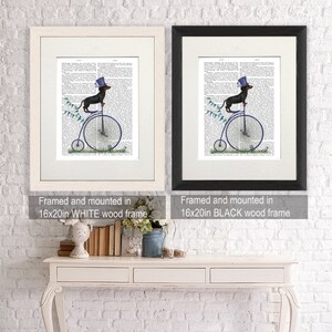 May include: A framed print of a dachshund wearing a top hat and riding a penny-farthing bicycle. The print is framed in a white wood frame and is 16 inches by 20 inches.