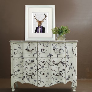 May include: A white dresser with a black floral design and three drawers. The dresser has a curved front and ornate legs. A framed print of a deer wearing a suit and bow tie is on top of the dresser.