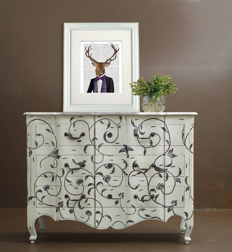 May include: A white dresser with a black floral design and three drawers. The dresser has a curved front and ornate legs. A framed print of a deer wearing a suit and bow tie is on top of the dresser.