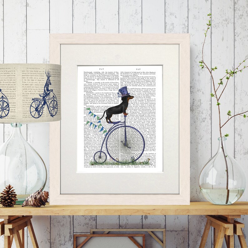 May include: A black and brown dachshund wearing a blue top hat rides a blue penny-farthing bicycle with blue and green flags on the handlebars. The image is printed on a page of a vintage book.