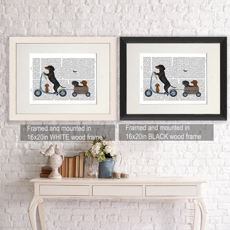 May include: A framed print of a black and brown dachshund riding a scooter with two smaller dachshunds in a wagon. The print is on a white background with black text. The frame is white and measures 16x20 inches.