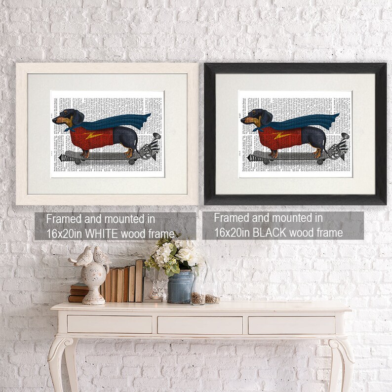 May include: A framed print of a dachshund wearing a superhero cape and riding a skateboard. The print is framed in a white wood frame and measures 16x20 inches.