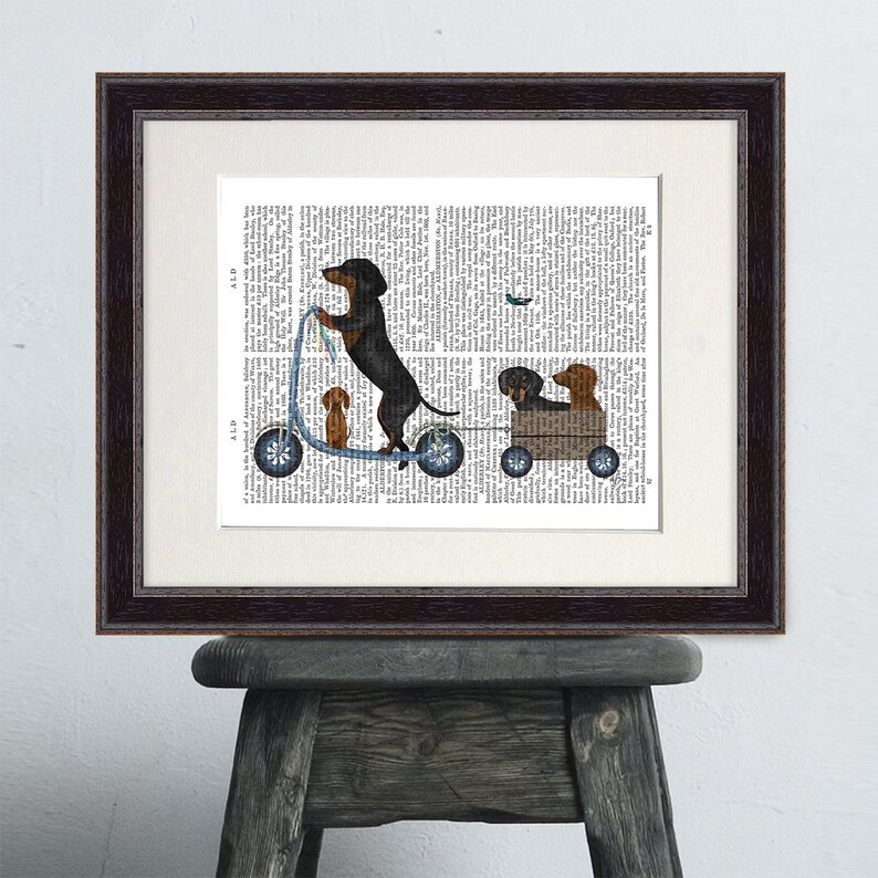 May include: A framed print of a black and brown dachshund riding a blue scooter with two other dachshunds in a wagon behind it. The print is on a white background with black text.