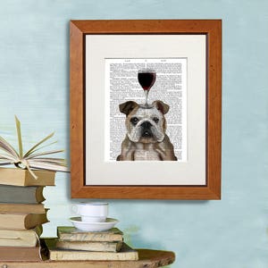 May include: A framed print of a bulldog with a glass of red wine on its head. The print is on a page of a vintage book with text. The frame is brown wood.
