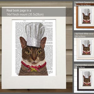 May include: A brown tabby cat wearing a white chef's hat and a red and white ribbon around its neck. The cat is printed on a book page with text. The image is framed in a black wood frame. The image is also available in white and caramel wood frames.