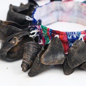 Puede incluir: Primer plano de un collar con cuentas oscuras en forma de cuerno. Las cuentas est&aacute;n ensartadas con una banda de tela estampada de colores rojo, azul, verde y blanco. El collar est&aacute; sobre un fondo blanco.